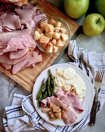 Top view of sliced ham and apple sauce on a cutting board with a dinner plate filled with ham, potatoes, apples, and green beans.
