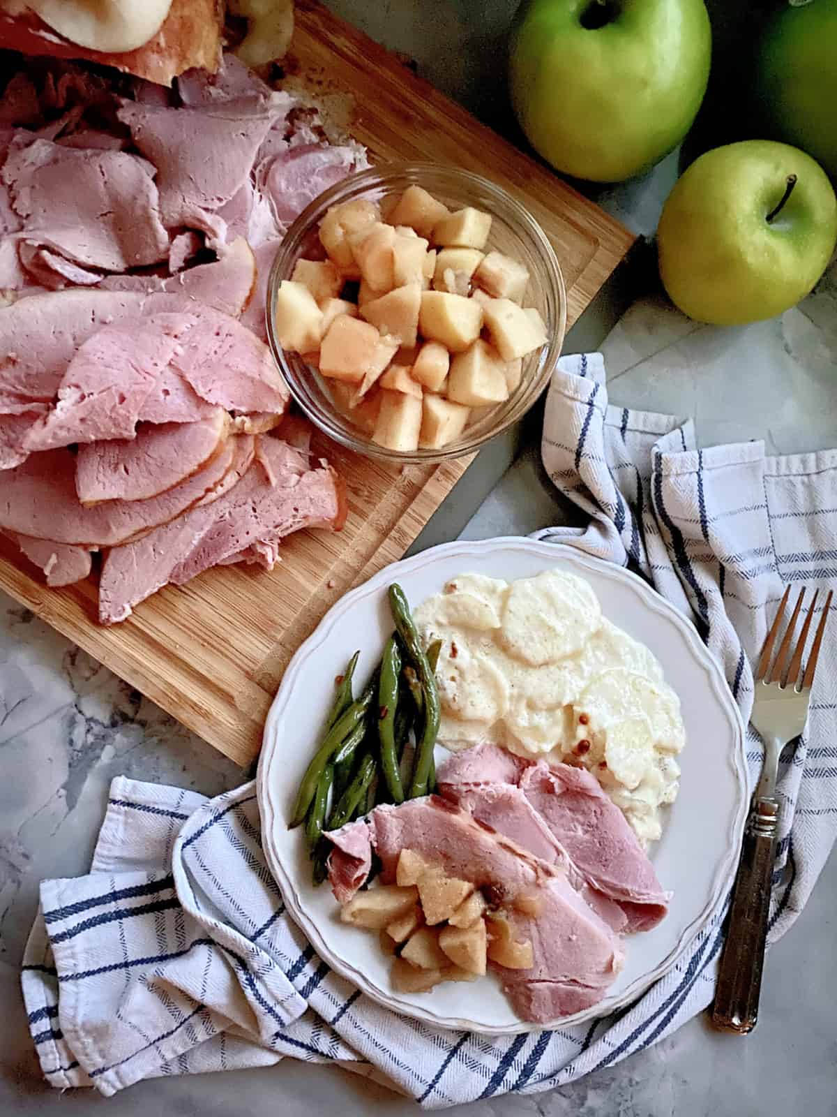 Top view of sliced ham and apple sauce on a cutting board with a dinner plate filled with ham, potatoes, apples, and green beans.