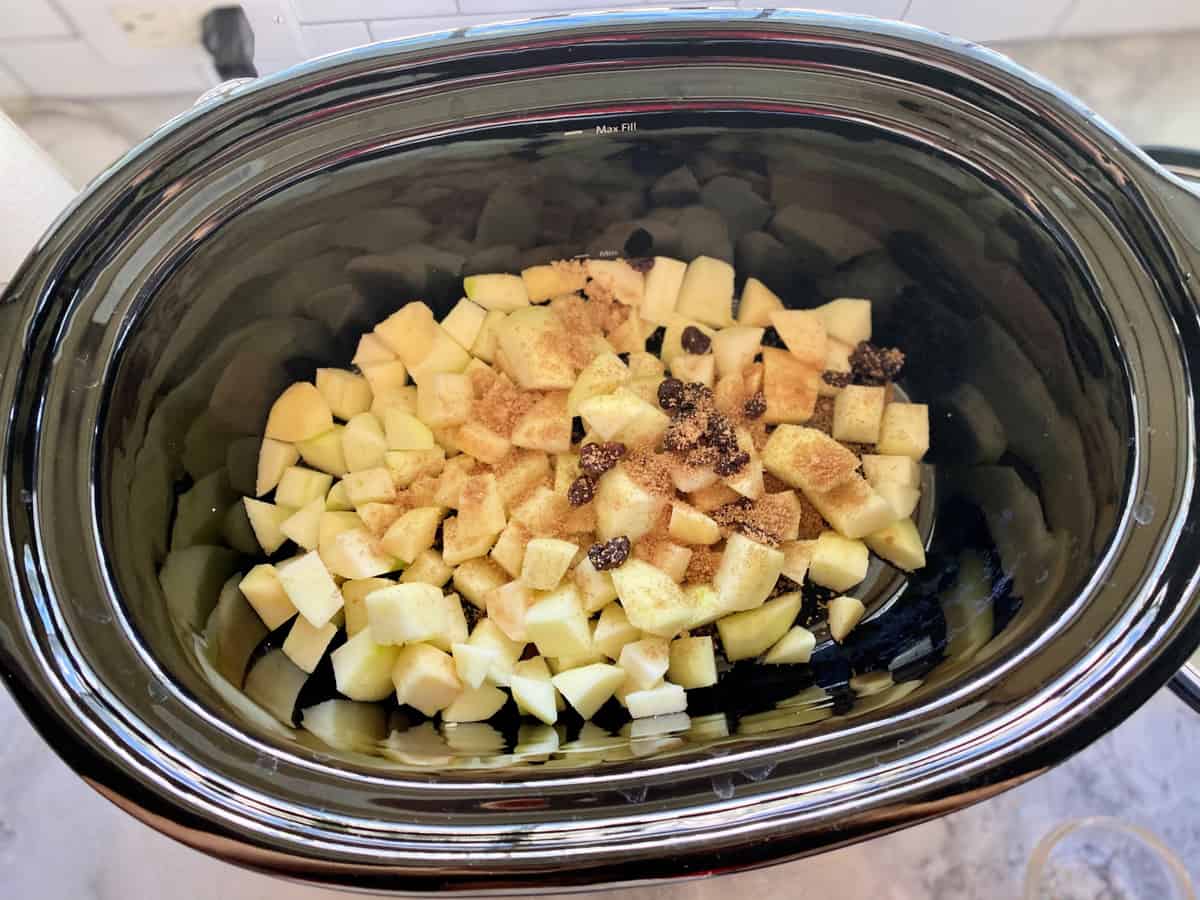 Top view of a black slow cooker filled with diced apples, spices, and raisins.