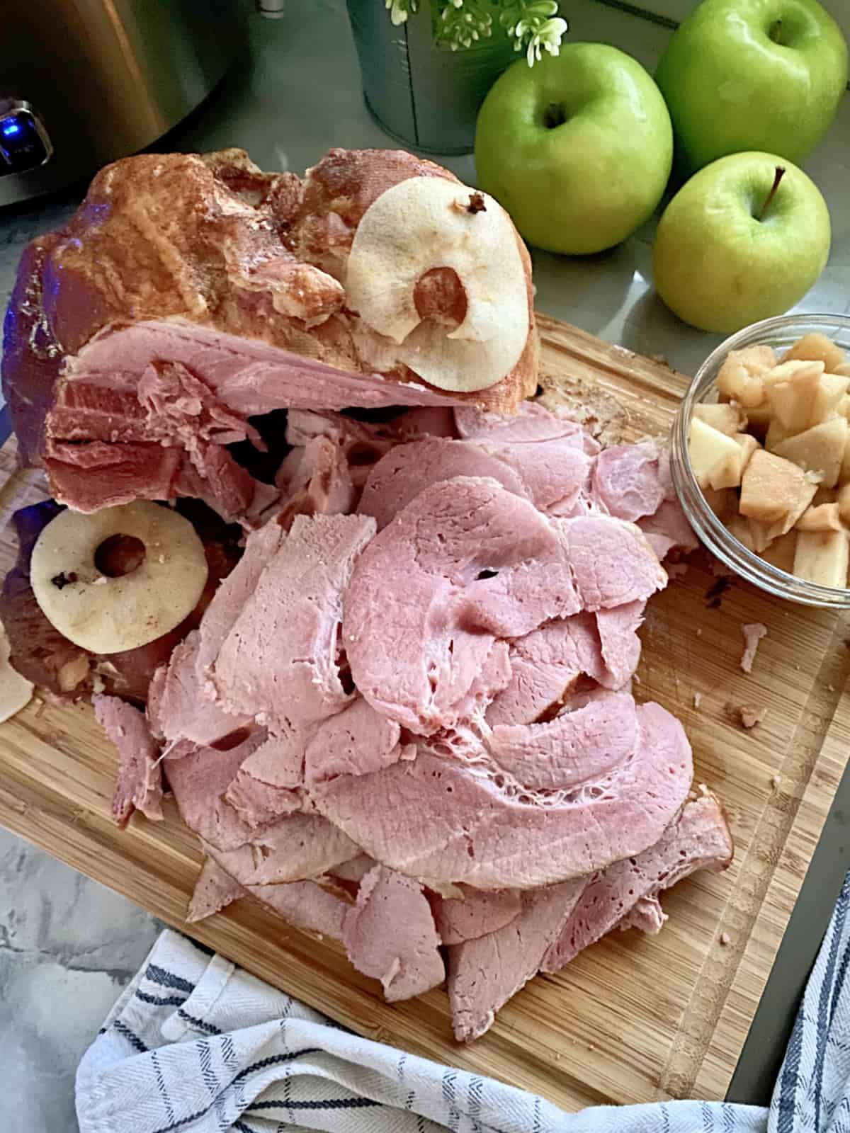 Top view of a wood cutting board with ham slices and a bowl of diced cooked apples next to it.