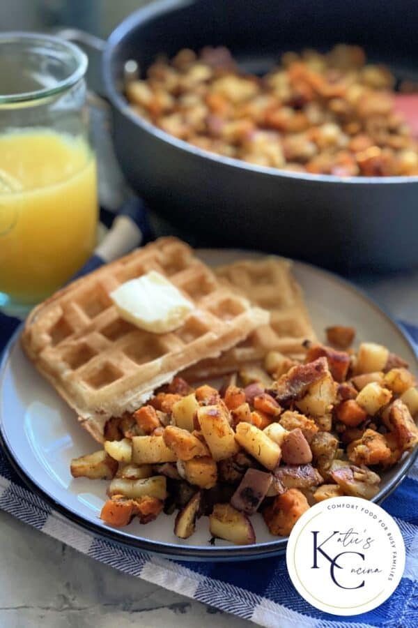 Plate filled with potatoes, waffles with orange juice and a skillet in the background.