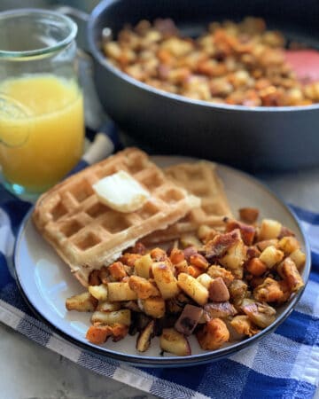 White plate with breakfast potatoes, waffles, and orange juice in the background with a skillet of potatoes.