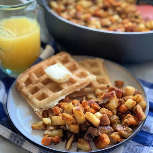 White plate with breakfast potatoes, waffles, and orange juice in the background with a skillet of potatoes.