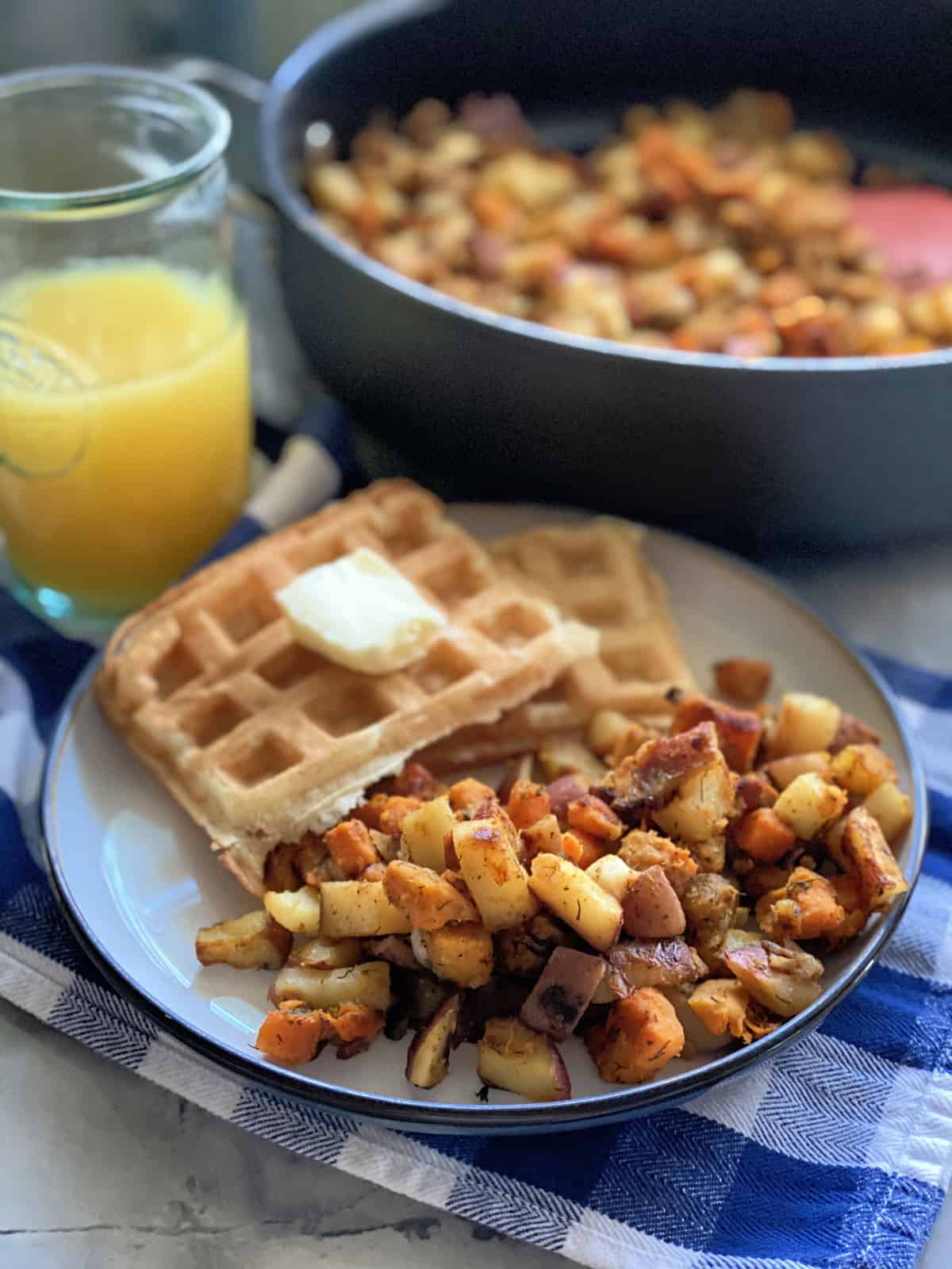 White plate with breakfast potatoes, waffles, and orange juice in the background with a skillet of potatoes.