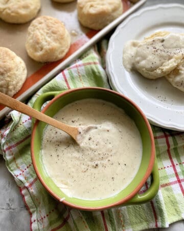 Top view of a green bowl filled with white gravy, plate of biscuits and gravy and a tray of biscuits.