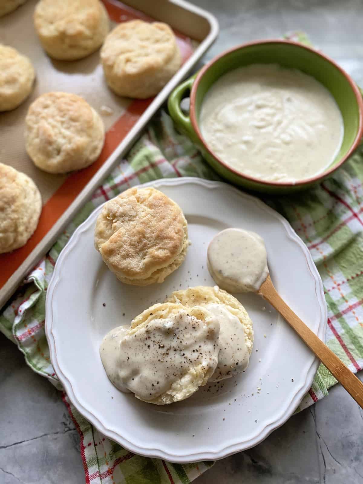 Top view of a white plate filled with biscuits and gravy with gravy and a tray of biscuits next to it.