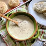 Top view of a green bowl filled with white gravy, and a tray of biscuits and a plate of biscuits and gravy next to it.