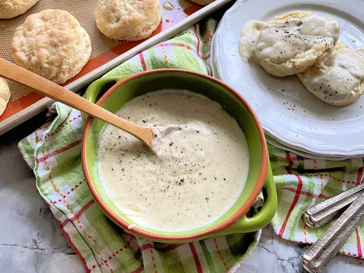 Top view of a green bowl filled with white gravy, and a tray of biscuits and a plate of biscuits and gravy next to it.
