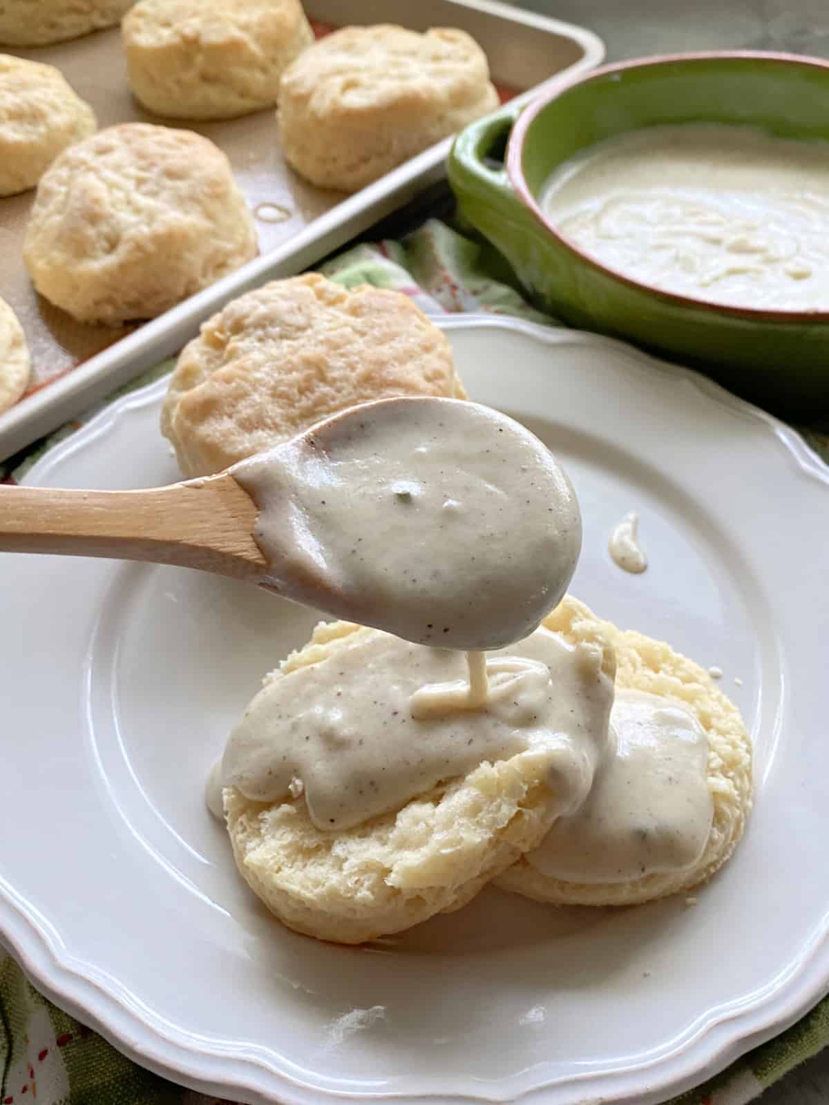 Wooden spoon drizzling white gravy on top of a biscuit.