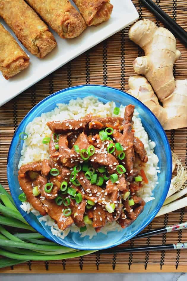 Top view of a blue bowl filled with white rice and beef on top with egg rolls and ginger on the side.