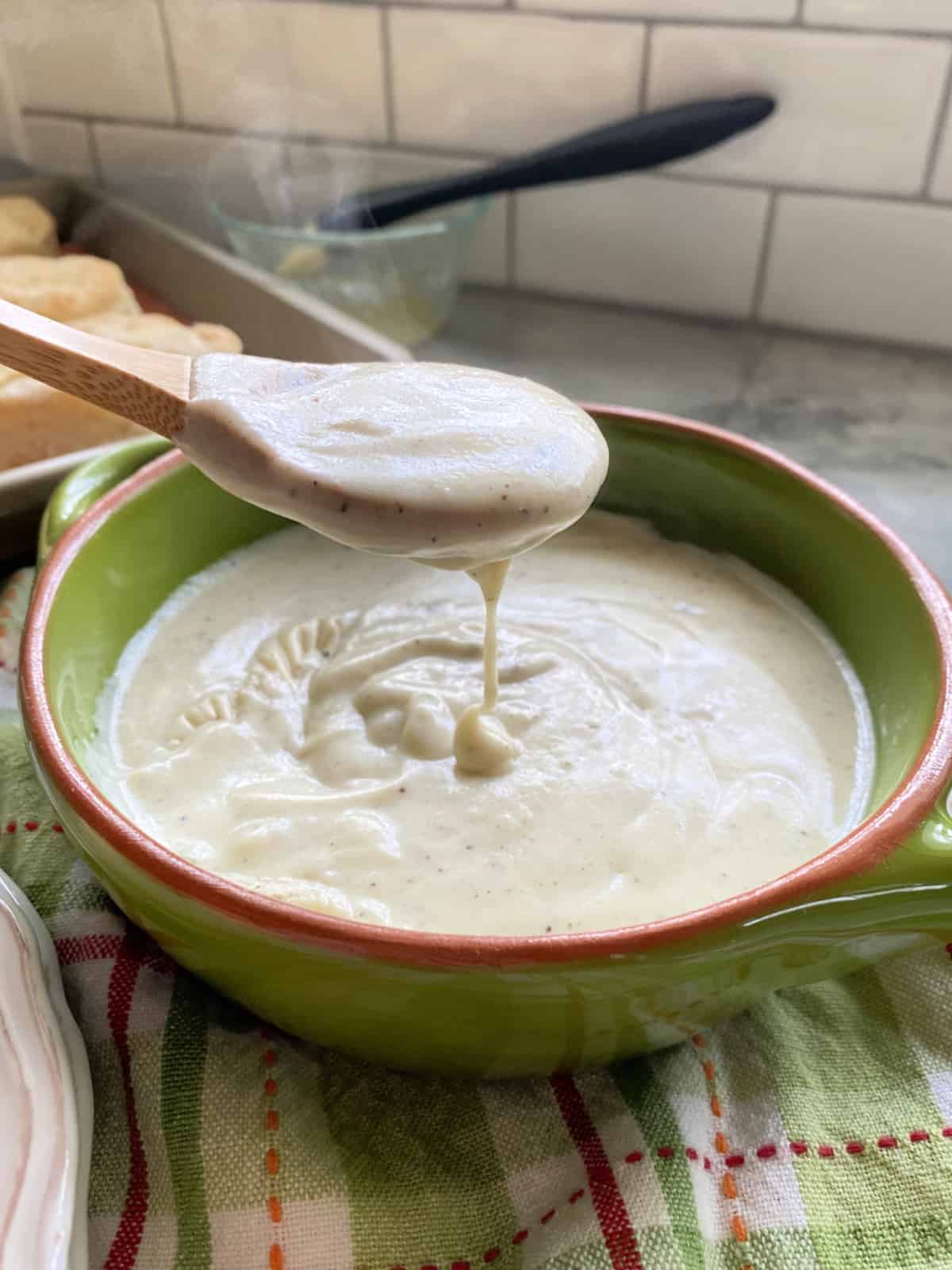 Wooden spoon drizzling white gravy into a green bowl full of white gravy.