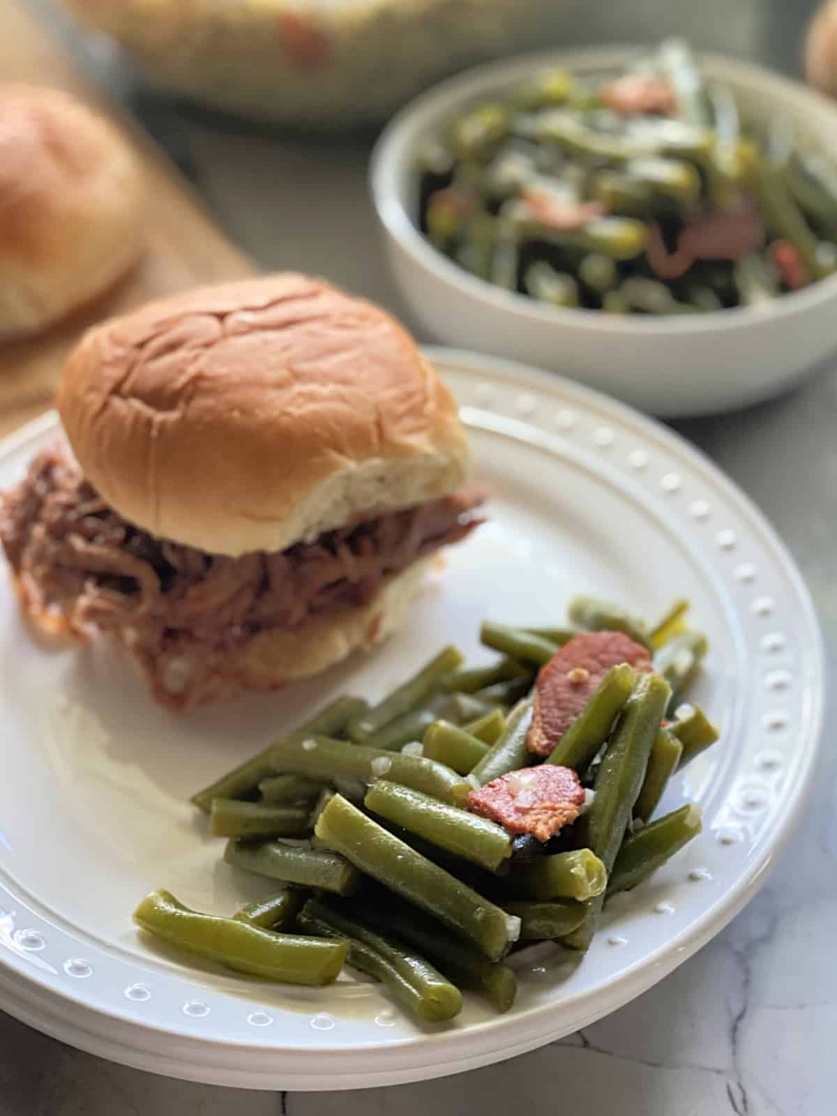 White dotted plate with green beans and bacon with a pulled pork sandwich sandwich in the background.