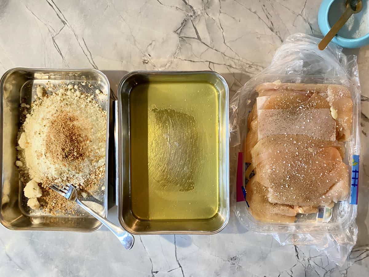 Top view of three rectangular trays with breadcrumbs, oil, and chicken cutlets.
