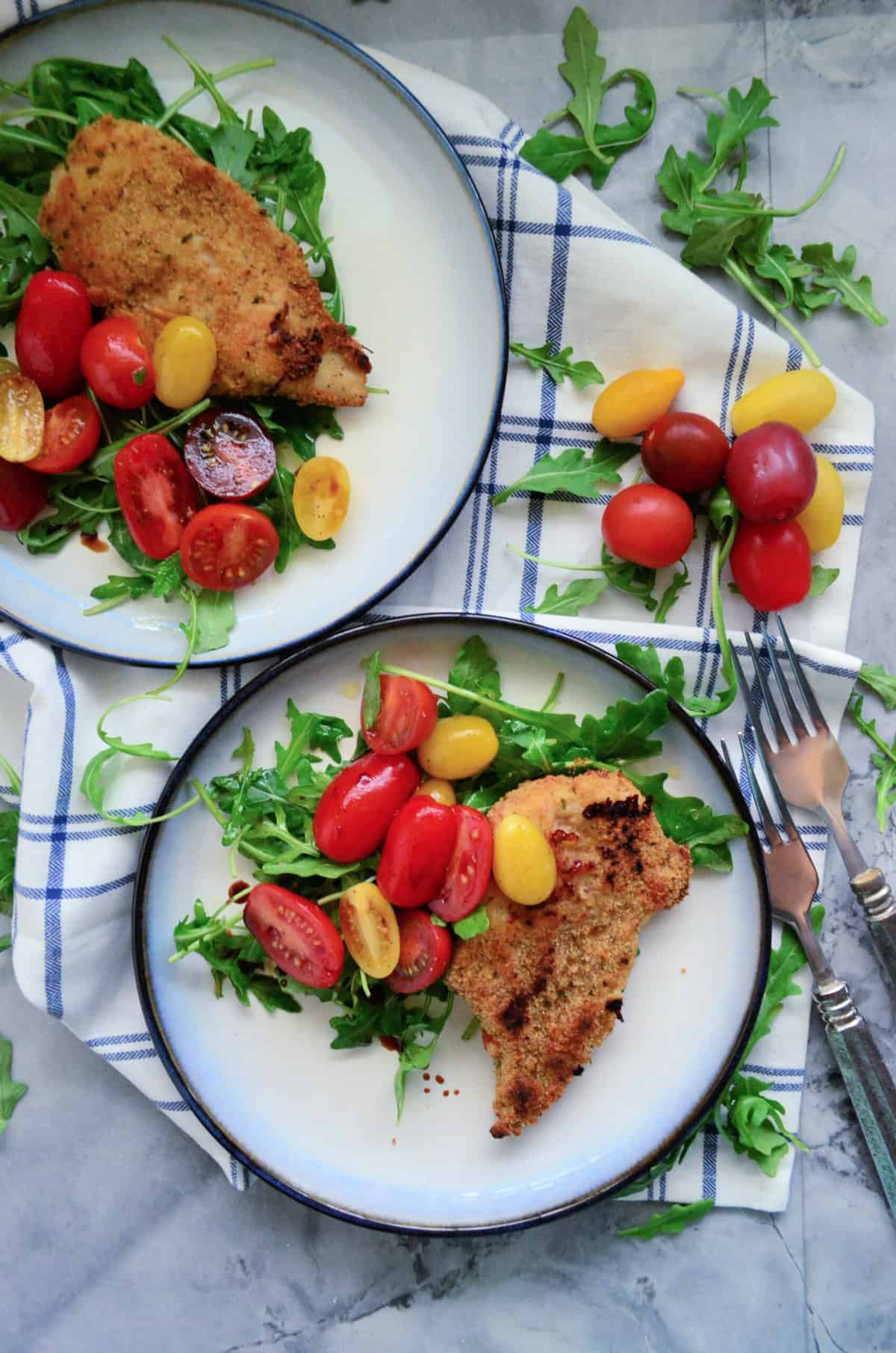 Top view of two white plate with a blue rim filled with arugula and tomato salad with a chicken cutlet on top.