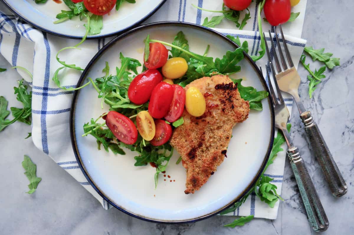 Top view of a white plate filled with an arugula and tomato salad with a chicken cutlet.