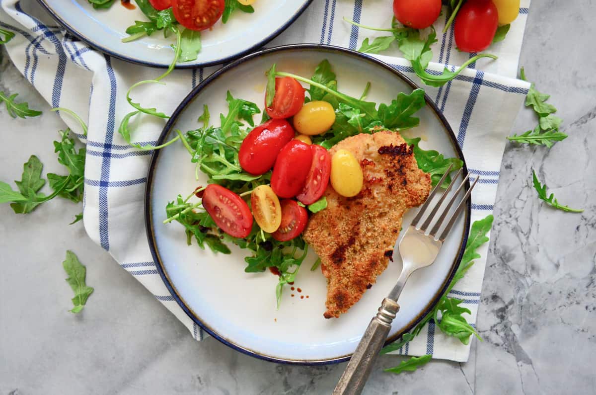 Top view of a white plate with a breaded chicken cutlet, arugula and tomato salad with a fork on the plate.