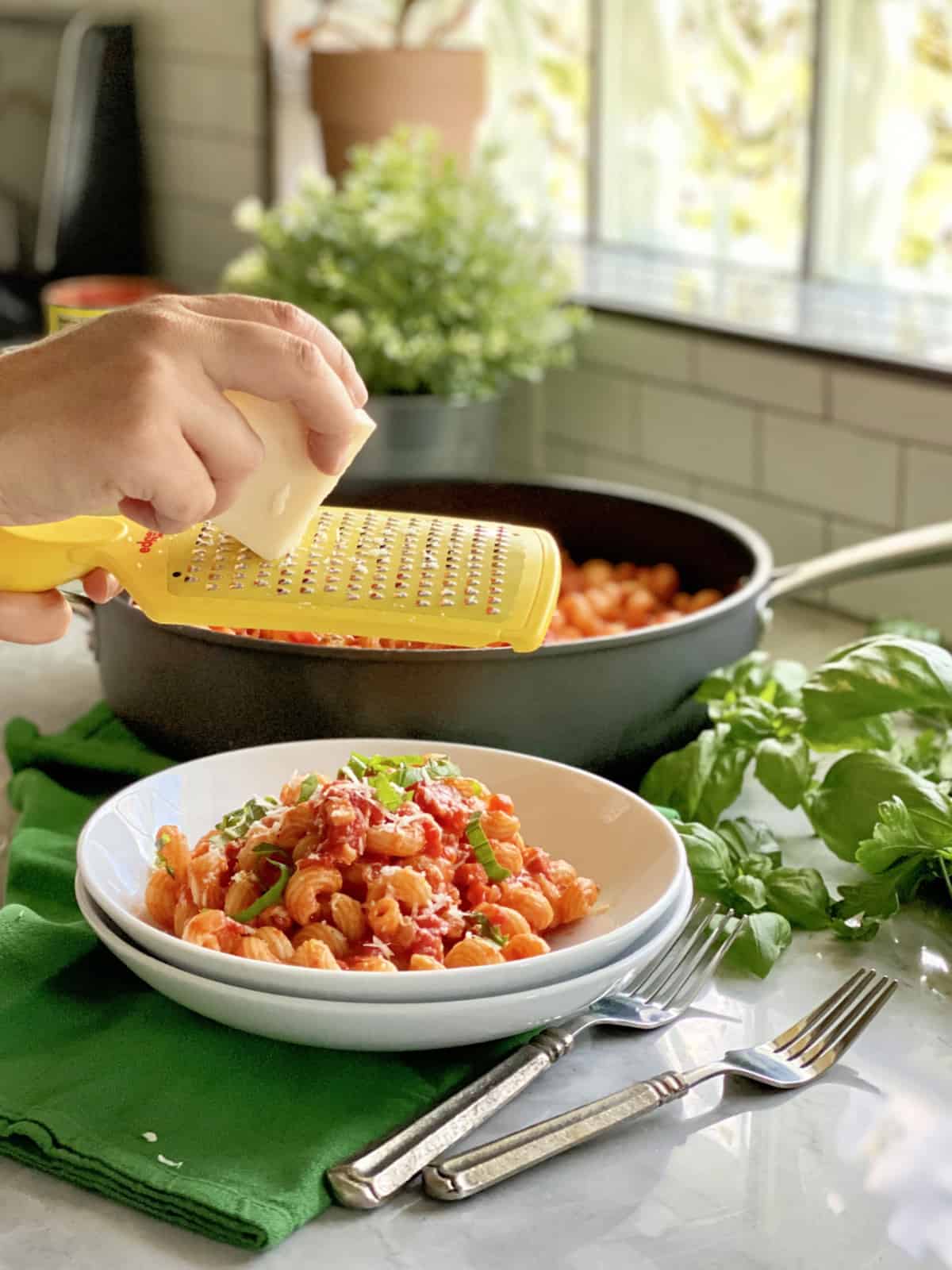 Female hand grating cheese over a bowl of pasta with skillet in the background.