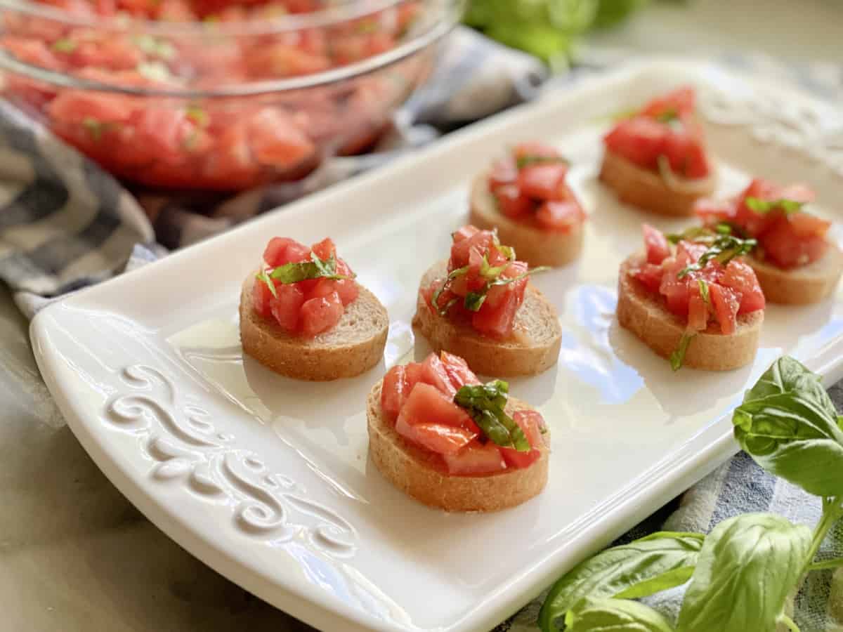 White platter filled with 7 pieces of baguette topped with bruschetta and a bowl of bruschetta in the background.