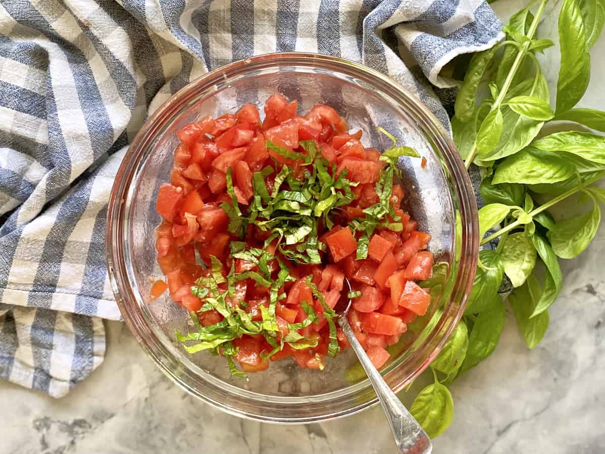 Top view of a glass bowl filled with diced tomatoes, basil threads, and a spoon.