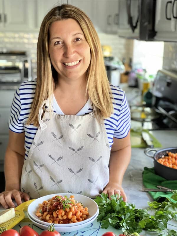 Female standing in a kitchen wearing an apron with a bowl of pasta and fresh herbs.