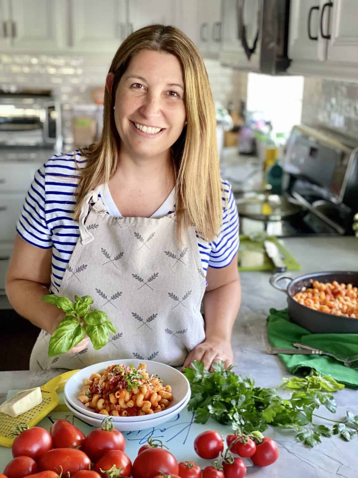 Female in apron holding basil with pasta and tomatoes on counter.