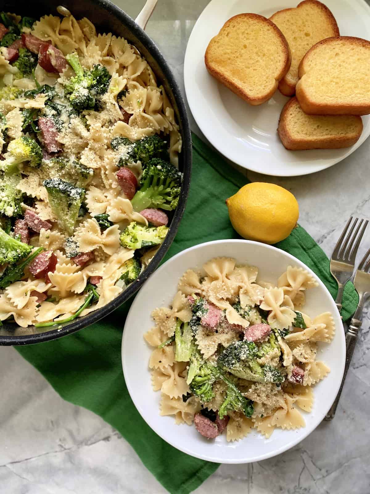 Top view of a black skillet and white bowl filled with bow tie pasta, broccoli, and kielbasa with bread on the side.