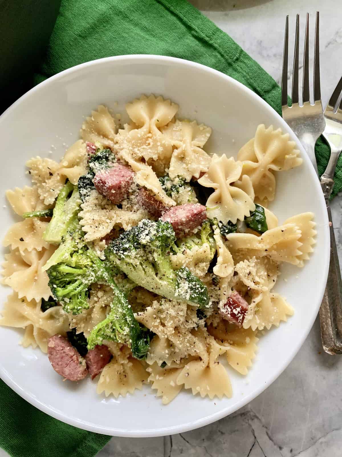 Top view of a white bowl filled with bow tie pasta, kielbasa, nad broccoli with forks on the side.