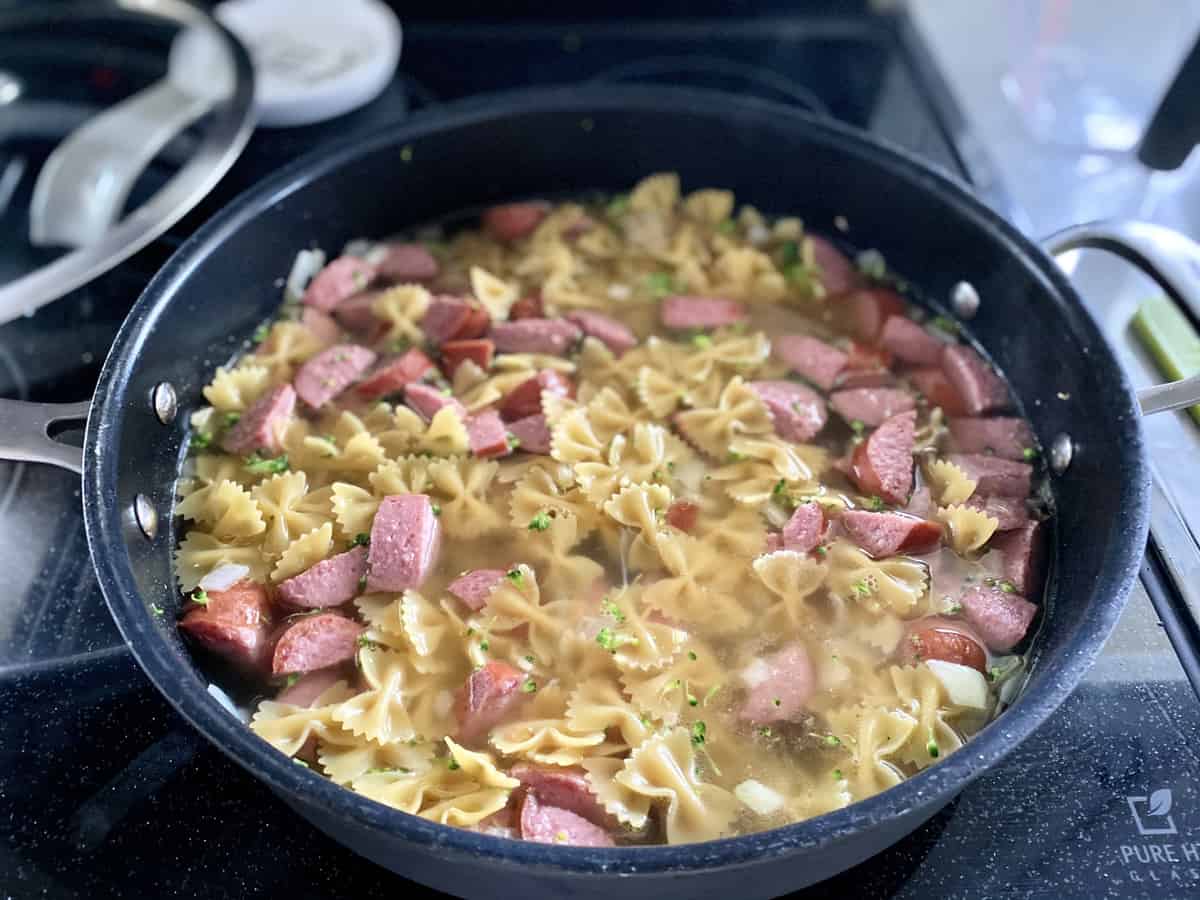 Black skillet filled with bow tie pasta, water and kielbasa.