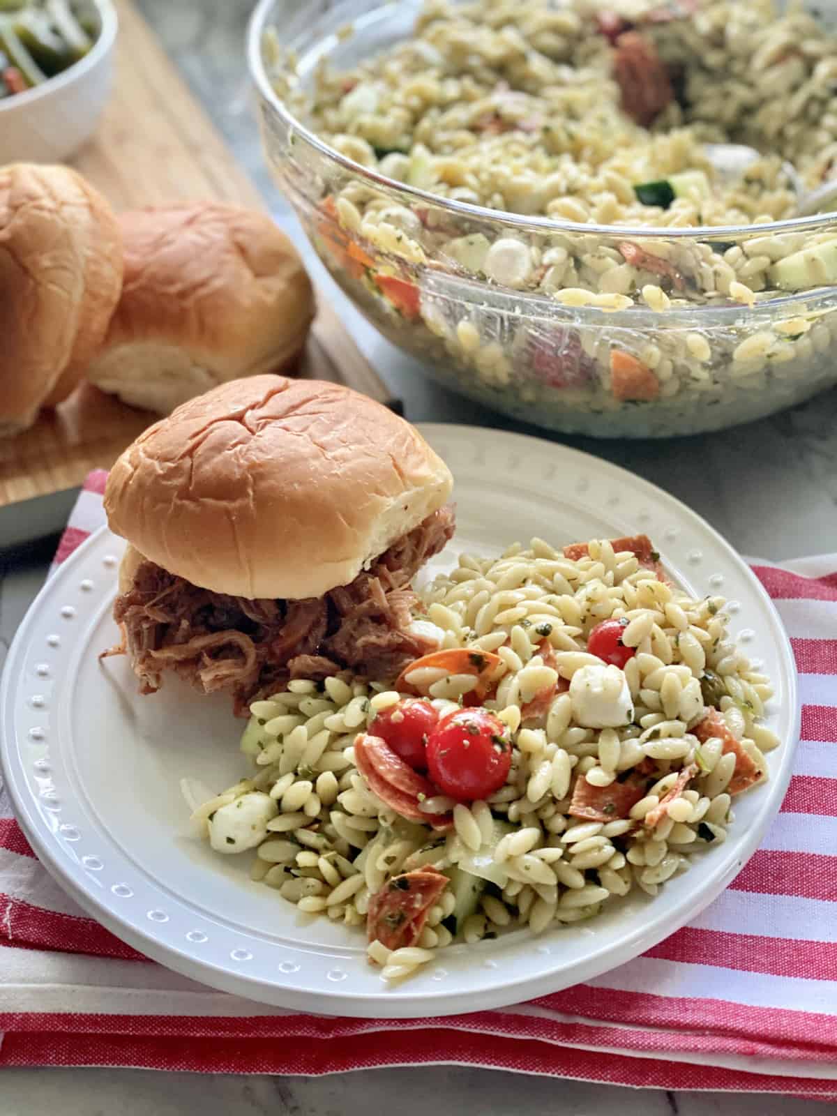 White plate filled with orzo pasta salad with pulled pork sandwich on the plate.