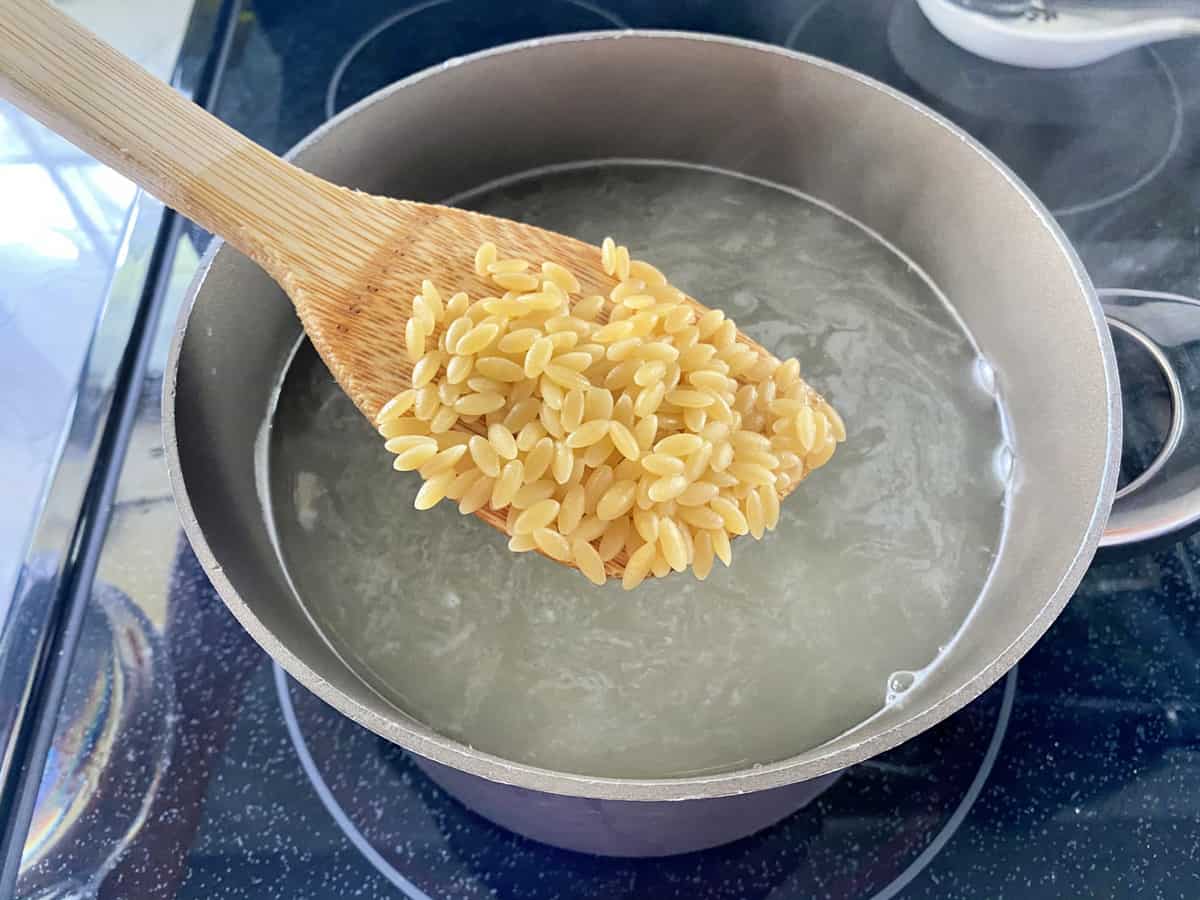 Wooden spoon showing orzo cooking a pot of boiling water on a stovetop.