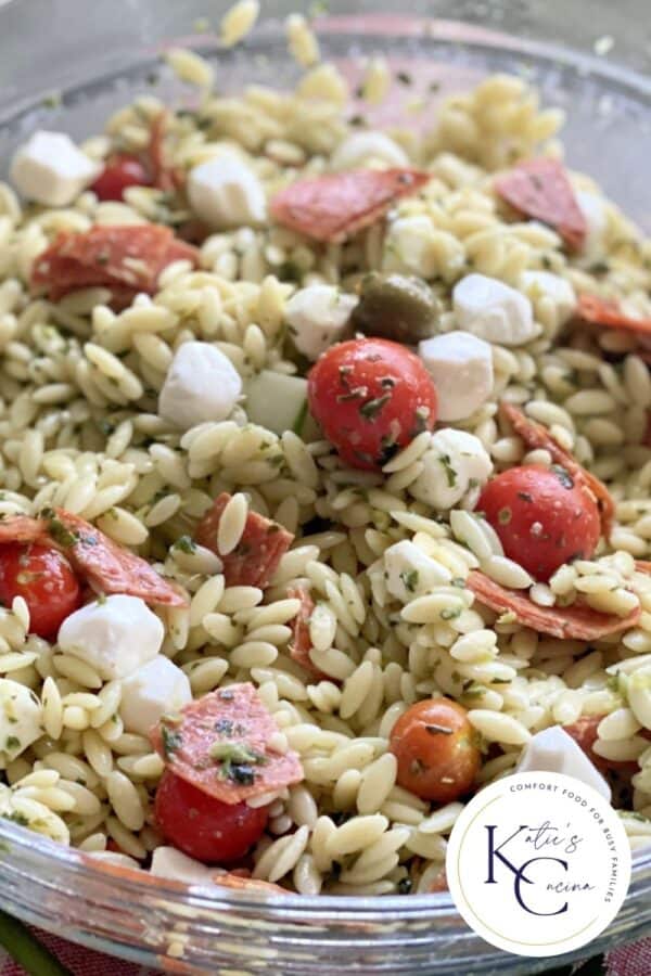 Close up of a glass bowl filled with Orzo Pasta Salad with logo on right corner.