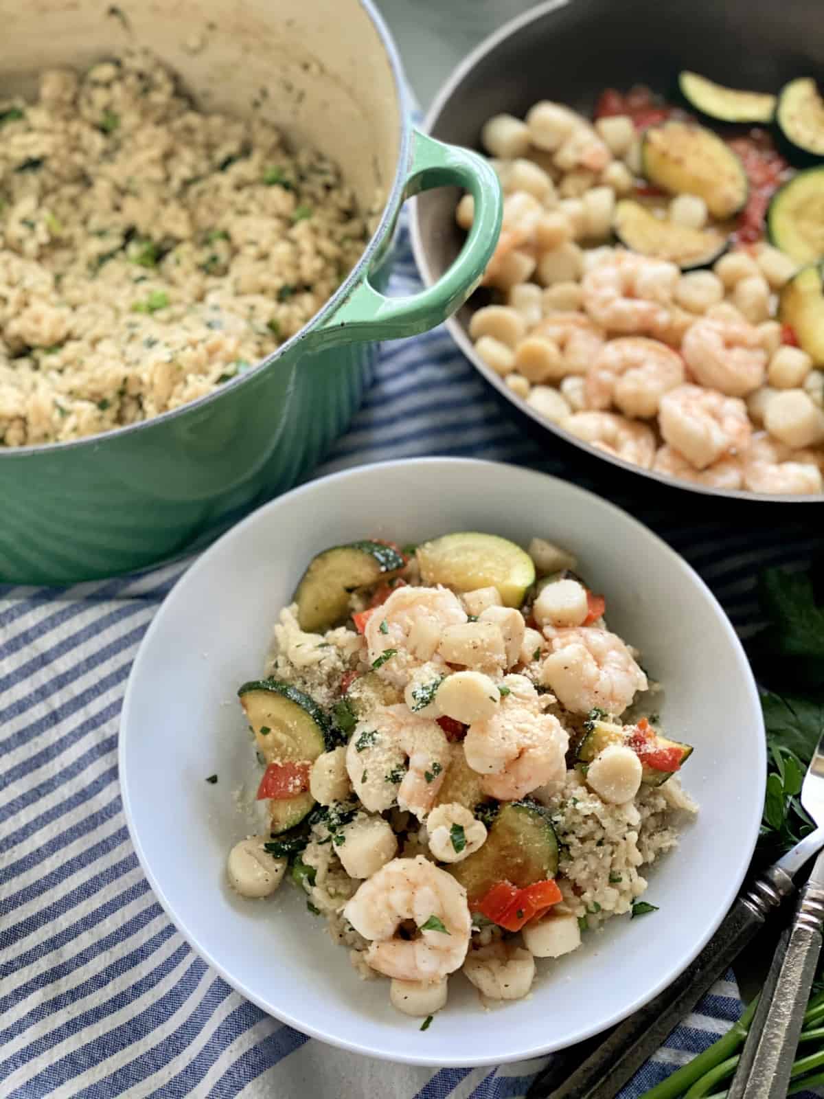 Top view of a white bowl filled with seafood risotto, green pot with risotto, and shrimp in a skillet.