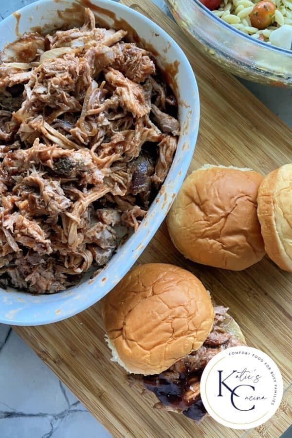 Top view of a bowl of pulled pork on a wood cutting board with buns and pulled pork sandwiches.