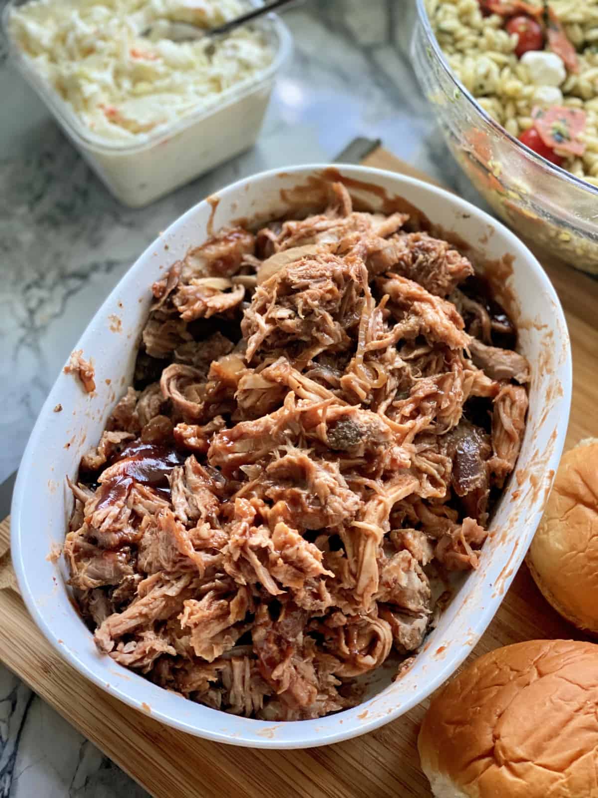 White oval baking dish with shredded pulled pork on a wood cutting board with sides around the dish.