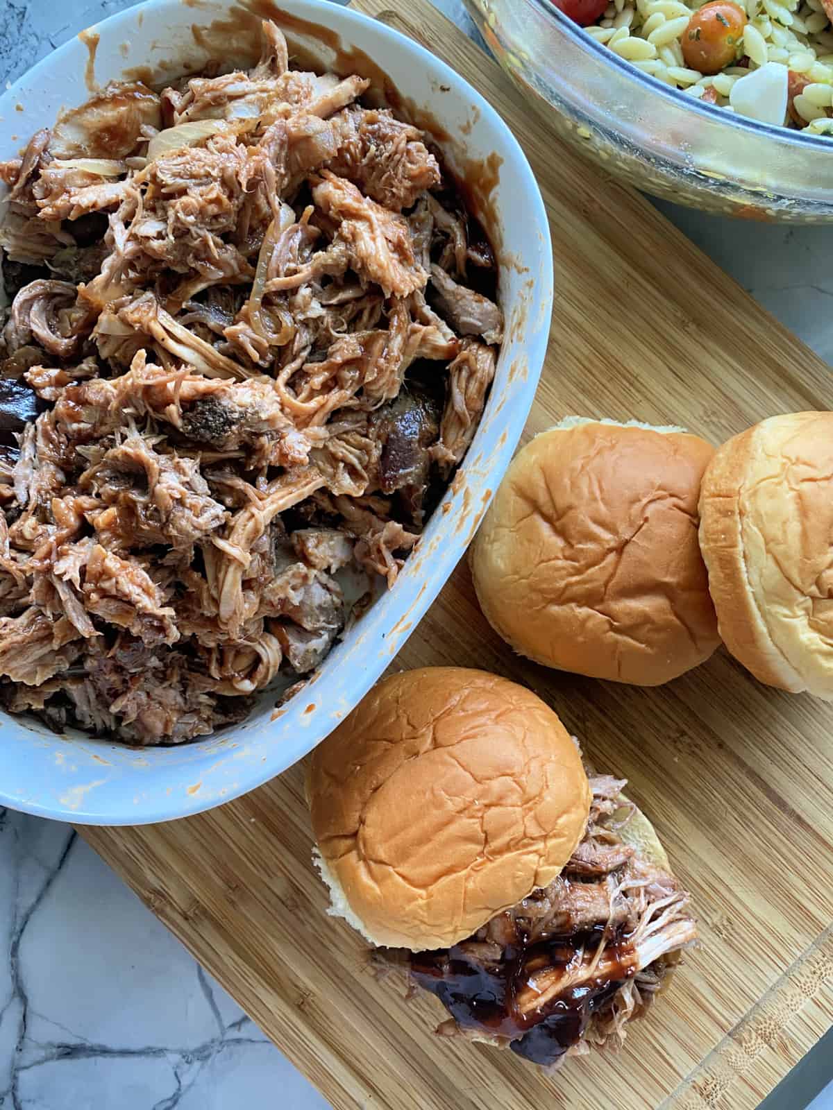 Top view of an oval dish on a wood cutting board with rolls and pulled pork sandwiches next to it.