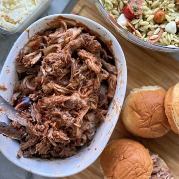 Top view of side dishes around a bowl of shredded pork with buns on cutting board.