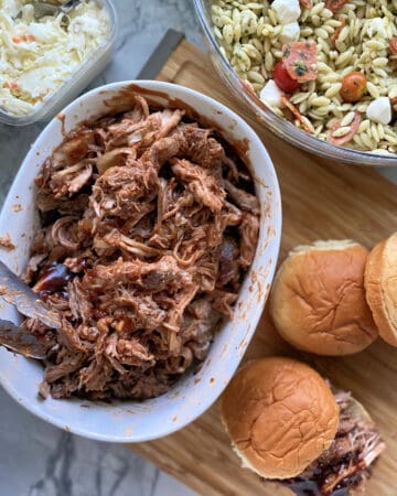 Top view of side dishes around a bowl of shredded pork with buns on cutting board.