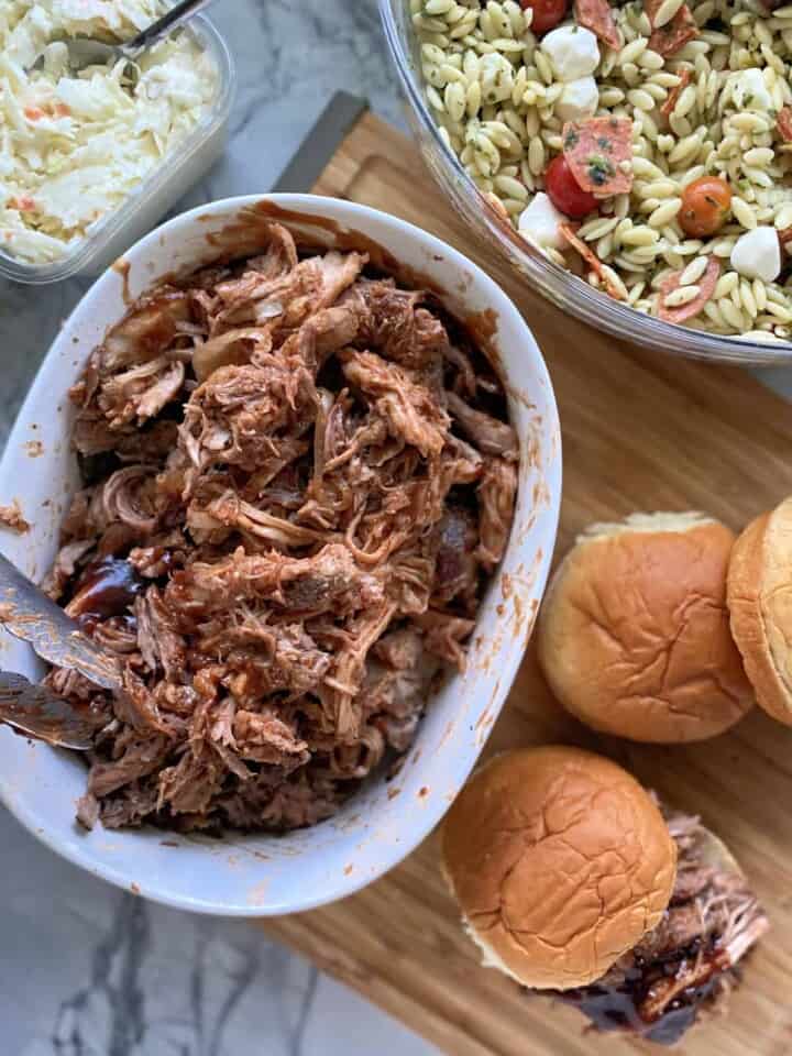 Top view of side dishes around a bowl of shredded pork with buns on cutting board.