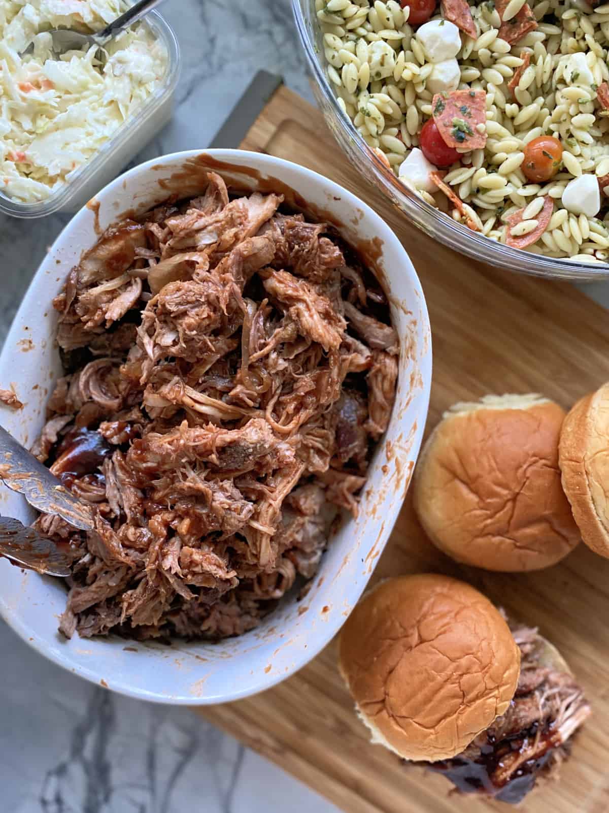 Top view of side dishes around a bowl of shredded pork with buns on cutting board.