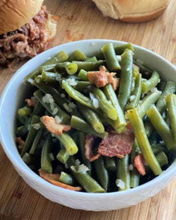 White bowl filled with chopped cooked green beans, onions, and bacon with pulled pork sandwiches in the background.