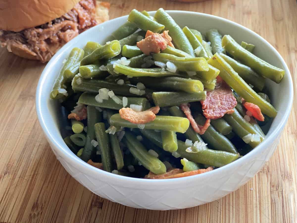 White bowl filled with green beans, onions, and bacon on a wood cutting board.