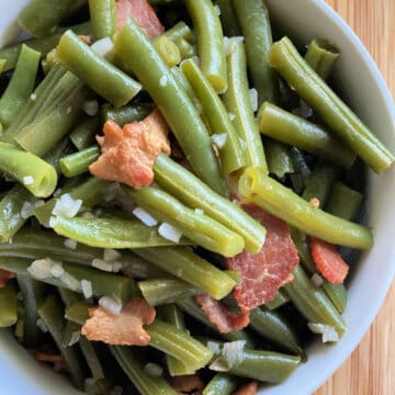 Top view of green beans with bacon and onions on a wood cutting board.