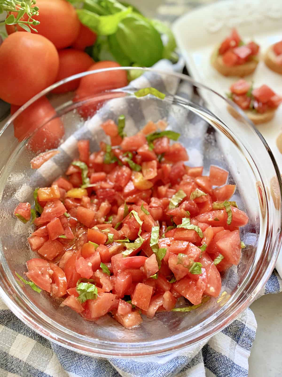 Top view of clear bowl with diced tomatoes with basil and tomatoes in background.