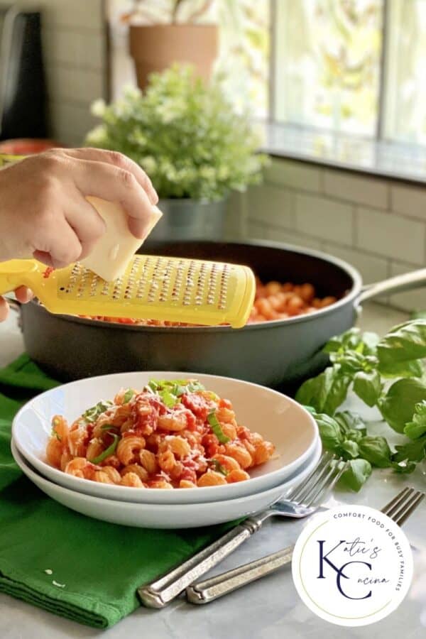 Female hand grating cheese over a bowl of red pasta with logo on right corner.