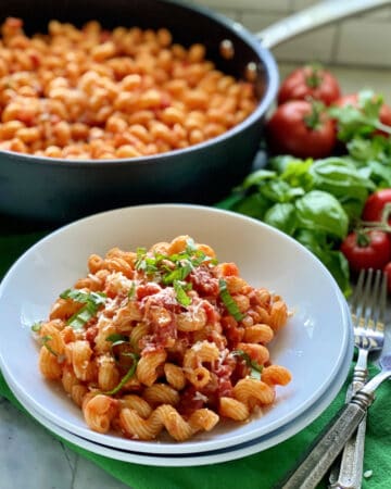 Two white bowls filled with corkscrew pasta topped with sauce, basil, and cheese with skillet in background.