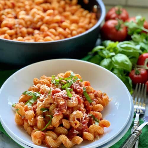 Two white bowls filled with corkscrew pasta topped with sauce, basil, and cheese with skillet in background.