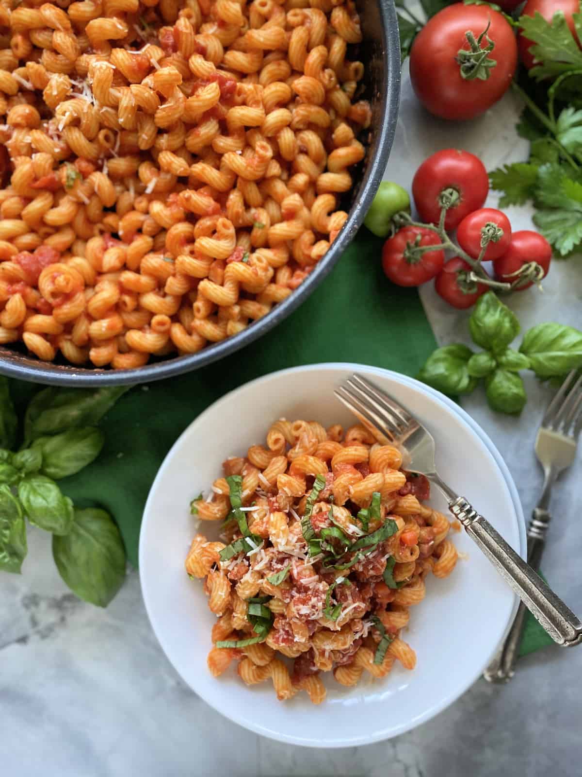 Top view of a skillet filled with pasta and white bowl filled with pasta, cheese, and basil.