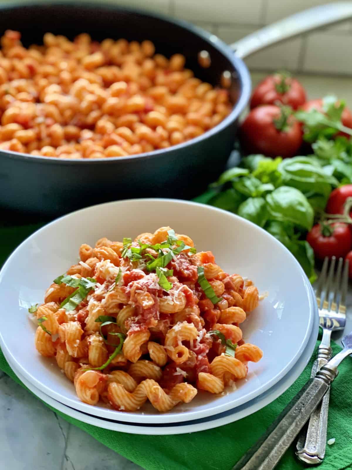 Two white bowls filled with corkscrew pasta topped with sauce, basil, and cheese with skillet in background.