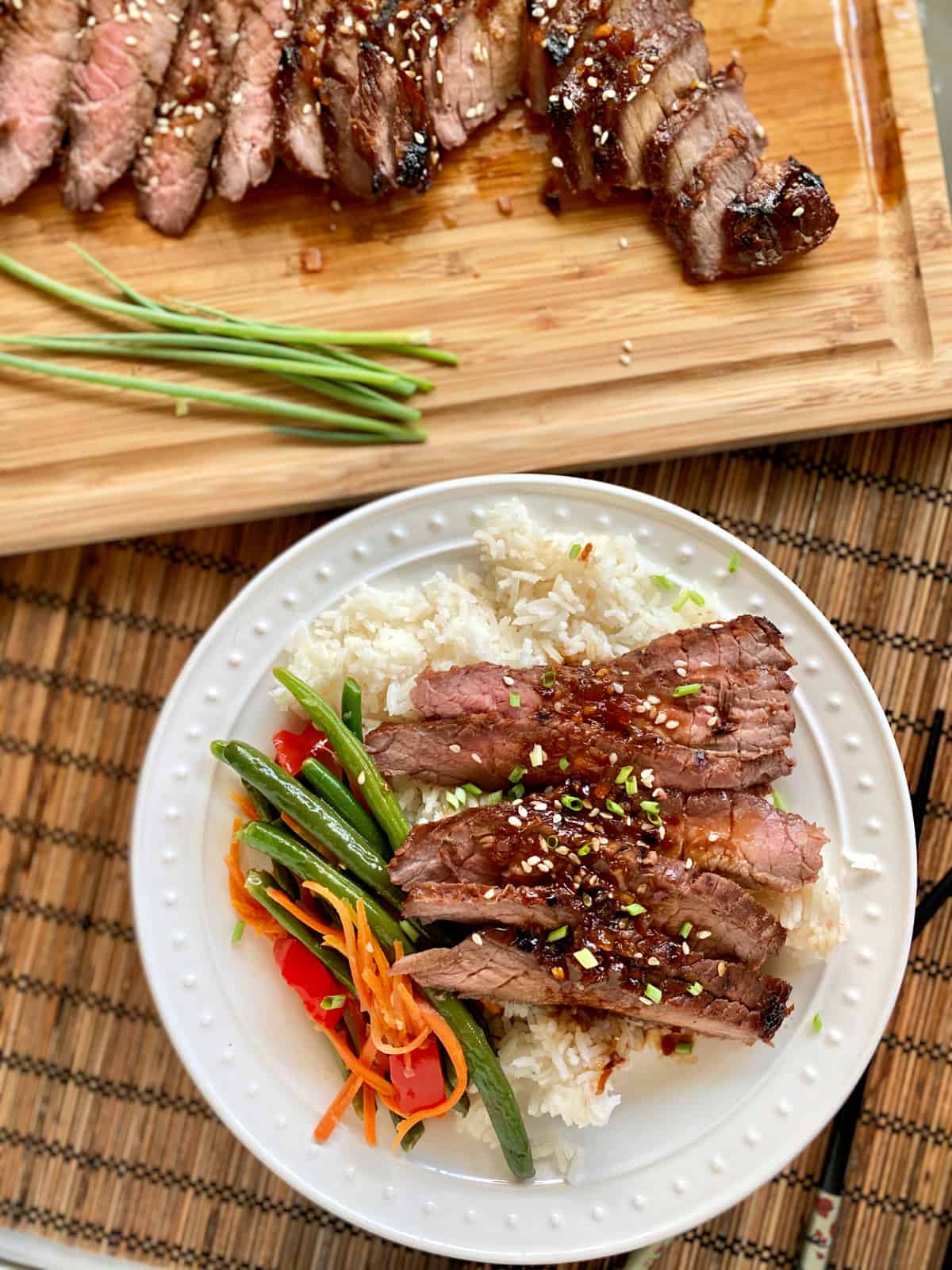 Top view of a white plate filled with slices of steak, rice, and green beans with a wood cutting board with more steak.