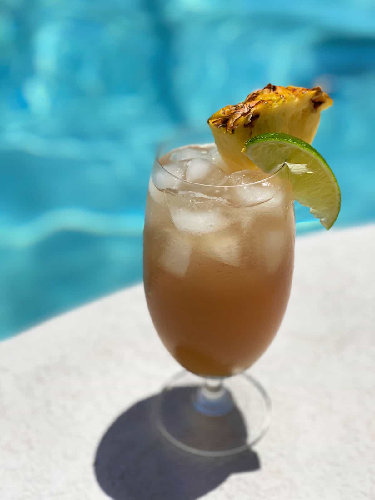 One glass of a cocktail next to a swimming pool garnished with a pineapple and lime.